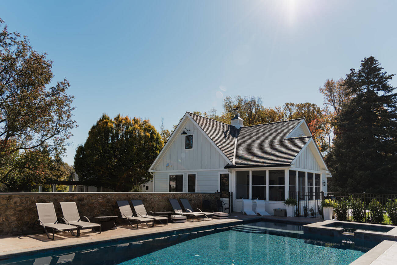 white pool house addition with cedar shake roofing and a cupola next to a large backyard swimming pool